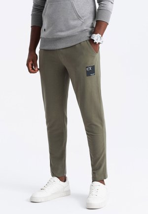 Olivegrüne Jogginghosen aus weichem Material mit schmaler Passform, ausgestattet mit einem schwarzen Logo-Patch auf der linken Tasche und einem elastischen Bund. Kombiniert mit weißen Sneakers.