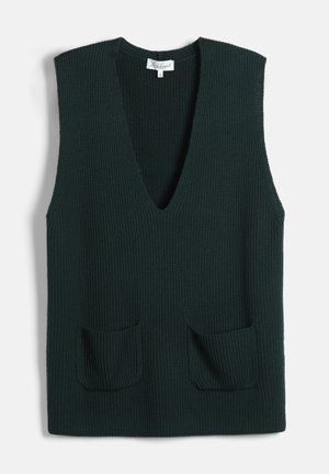 Vrouw draagt een donkergroene geplooide rok met split, bijpassend gebreid vest, wit overhemd, groene handschoenen, zwarte laarzen en zwarte schoudertas op een witte achtergrond.