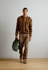 Multikleurige gebreide trui met textuurlijnen, bruine broek en beige schoenen. Houdt een groene gewatteerde tas vast. Houten vloeren op de achtergrond.