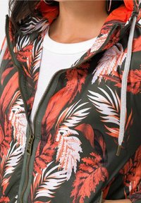 Vihreäpohjainen huppari, jossa on eloisat punaiset ja valkoiset lehtikuvioinnit. Metallivetoketju ja vaaleanharmaat kiristysnauhat korostavat mallia.