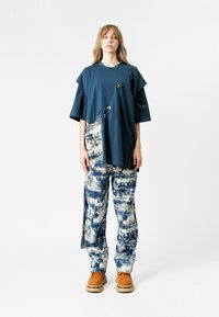 Una joven se encuentra de pie vistiendo una camiseta azul marino de gran tamaño con pantalones de estampado abstracto en blanco y azul marino, y zapatos de plataforma beige, frente a un fondo blanco.