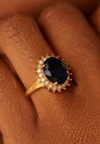 Gouden ring met een grote ovale blauwe edelsteen, omringd door kleine witte stenen, met een gestructureerde band en sierlijke zettingen.