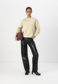 Sudadera beige, pantalones de cuero anchos negros y botas de tacón grueso negras. La modelo sostiene un bolso burdeos, mostrando un diseño limpio y moderno.