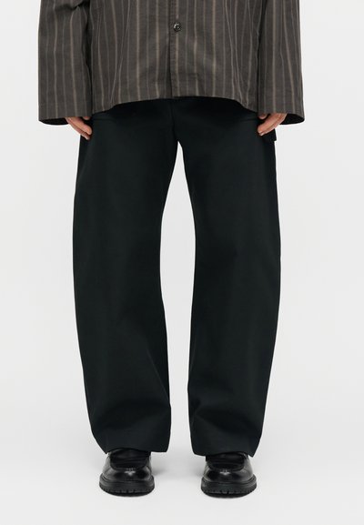 Άτομο που φοράει φαρδιά μαύρα παντελόνια, μαύρα δερμάτινα παπούτσια και καφέ πουκάμισο με ρίγες, στέκεται μπροστά σε απλό φόντο.