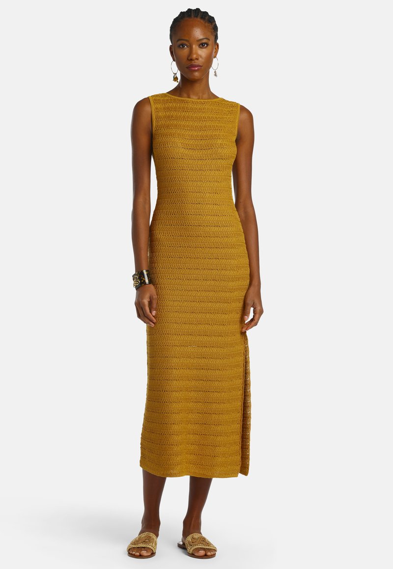 Femme debout portant une robe midi tricotée sans manches jaune moutarde avec une fente sur le côté, associée à des sandales tressées, des boucles d'oreilles créoles et un bracelet manchette.