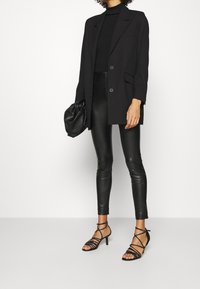 Kvinna klädd i svart kavaj, polotröja, läderbyxor, sandaler med remmar, hållande en svart rynkad clutchväska, stående på vit bakgrund.