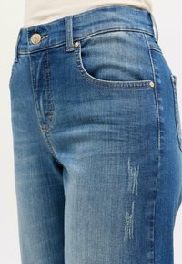 Jean en denim avec une taille mi-haute, dégradé en tons de bleu, quelques détails usés et des accessoires en métal doré sur les poches.