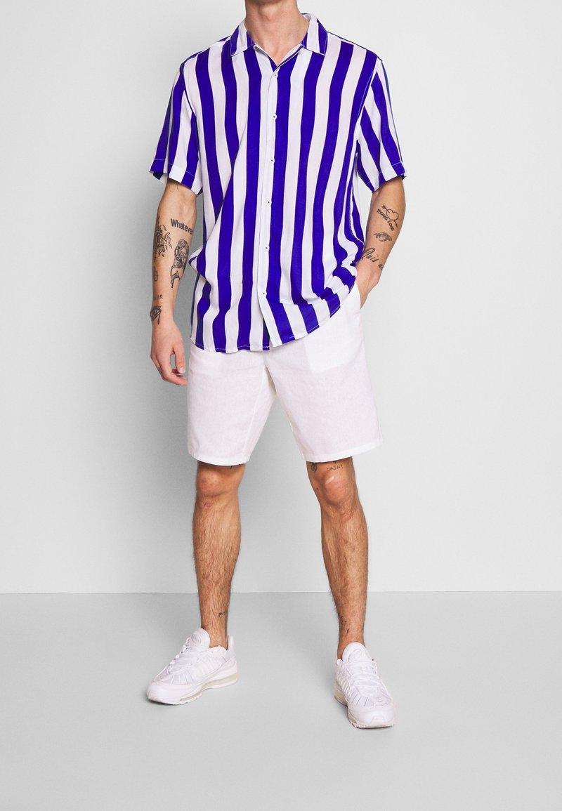 Homme portant une chemise à manches courtes rayée verticalement bleue et blanche, un short blanc, des baskets blanches, debout avec une main dans la poche sur un fond uni.