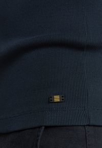 Marineblauwe geribbelde shirtstof met een zwart label met drie gele horizontale strepen aan de onderkant, die textuur en ontwerp accentueren.
