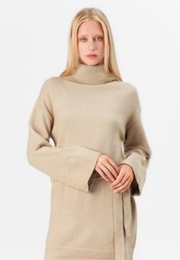 Beige polotröja i mjuk knit, med vida ärmar och en knytdetalj i midjan. Slät textur med ribbad fåll.