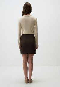 Beige lange mouw gebreide top, met textuur, gecombineerd met een donkerbruine rok met zakken. Model staat met blote benen en lichte schoenen.