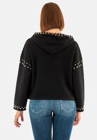Sudadera negra corta con tachuelas plateadas en las mangas y la capucha. Hecha de una tela suave, presenta un corte relajado y mangas largas. Combinada con jeans azules.