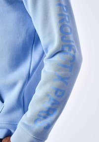 Sudadera azul claro con una textura suave que presenta "PROJECT X PARIS" impreso en un azul más oscuro a lo largo de la manga izquierda.