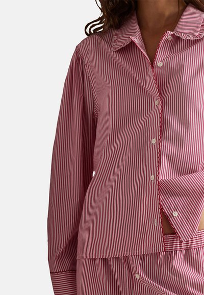 Femme portant un pyjama à boutons à manches longues à rayures verticales rouges et blanches, avec un pantalon assorti, sur fond uni.