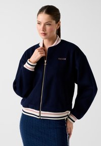 Blouson aviateur en polaire bleu marine avec poignets et ourlet côtelés rayés blancs et roses. Dispose d'une fermeture éclair et d'un logo brodé "GUESS".