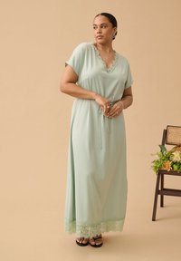 Femme portant une robe longue vert clair avec une bordure en dentelle au niveau de l'encolure et de l'ourlet, debout, les mains jointes, à côté d'une chaise avec des fleurs.