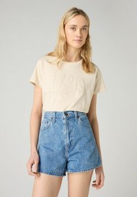 Beige t-paita, jossa on kohokuvioitu "LEVI'S" -logo, yhdistettynä korkeavyötäröisiin sinisiin farkkushortseihin, joissa on klassinen viiden taskun muotoilu.