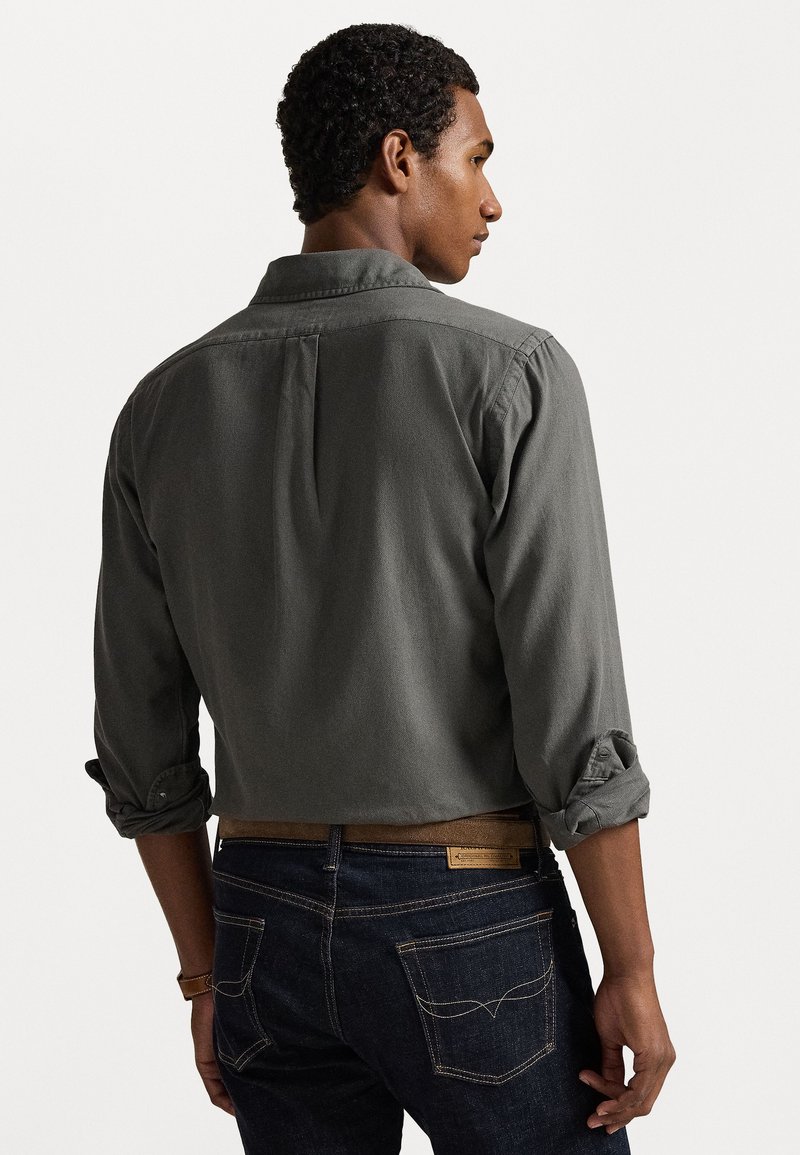 Graues Baumwollhemd mit hochgerollten Ärmeln, Rückenteil mit Detail, kombiniert mit dunklen Denimjeans mit sichtbaren Nähten und aufgesetzten Taschen.