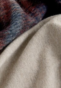 Puha gyapjú anyag bézs színben, texturált felülettel; rétegezve egy bolyhos, színes kötött anyaggal, amely kék, piros és szürke árnyalatait tartalmazza.