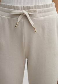 Beige Jogginghose mit rippierter Taille, verstellbarem Kordelzug und goldfarbigen Spitzen, ausgestattet mit einer weichen Textur und einem schlichten Design.