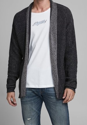 Homme portant un cardigan gris foncé ouvert sur un t-shirt graphique blanc et un jean bleu déchiré, se tenant devant un fond uni.