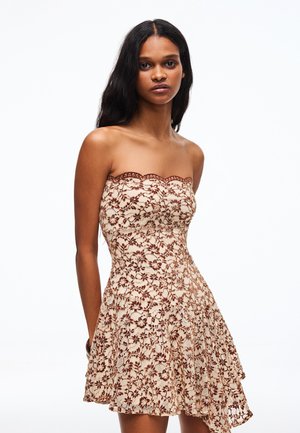 Frau mit langen dunklen Haaren, die ein trägerloses beiges Kleid mit brauner Blumen-Spitze trägt, steht vor weißem Hintergrund.