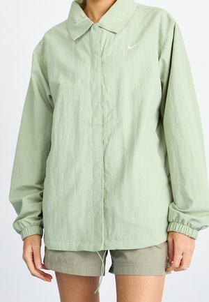 Osoba nosząca jasnozieloną kurtkę z długim rękawem z elastycznymi mankietami oraz beżowe szorty na jednolitym tle.