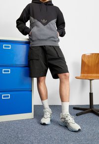 Person som bär en svart och grå hoodie, svarta cargoshorts, vita strumpor och grå sneakers, stående vid ett blått arkivskåp och en trästol.