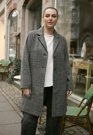 Grau karierten Wollmantel mit Reverskragen und Knopfschluss, kombiniert mit einem strukturierten weißen Oberteil und schwarzen Hosen, im Freien aufgestellt.