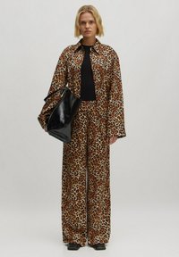 Leeuwenshirt met een panterprint en wijde broek in bruin en tan, gemaakt van gladde stof. Zwarte tas met een glanzende afwerking, gedragen op de schouder.