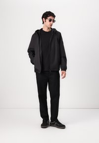 Sort letvægtsjakke med fast hætte, parret med en sort t-shirt og bukser. Modellen bærer solbriller og sorte sneakers.
