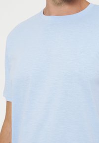 Κοντινό πλάνο ενός ατόμου που φοράει ανοιχτό μπλε μπλουζάκι με στρογγυλή λαιμόκοψη και μικρό κεντημένο "P" στο αριστερό στήθος.