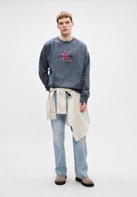 Sweatshirt cinza com um logótipo bordado ao centro, jeans azuis e uma camisa riscada com botões atada à cintura. Botas castanhas até ao tornozelo.