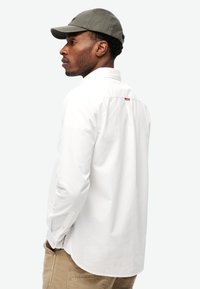 Camisa blanca de manga larga con botones y un pliegue en la espalda, combinada con una gorra de béisbol verde oscuro y pantalones caqui. Detalles visibles de los botones y el cuello.