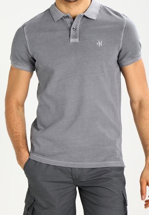 Mann trägt ein eng anliegendes graues Poloshirt mit kurzen Ärmeln und kleinem weißem besticktem Logo auf der Brust und dunkelgraue Cargoshorts.