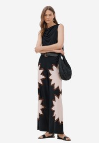 Maglia nera con cappuccio abbinata a pantaloni a gamba larga con una base crema e un motivo a raggiera in nero e arancione. Borsa intrecciata nera e sandali.