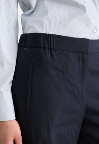 Calças azul marinho com cintura elástica, apresentando um bolso lateral e um tecido suave e texturizado. Combinadas com uma camisa de botão azul clara.