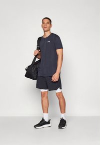 Muž v černém sportovním oblečení a teniskách, nesoucí černou sportovní tašku přes rameno, stojící před jednobarevným bílým pozadím.