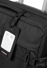 Bolsa de viaje negra con una tela texturizada, que cuenta con una etiqueta de identificación transparente, cierre de hebilla y un asa resistente para un transporte conveniente.