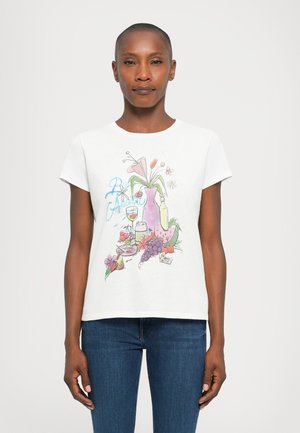 Woman wearing a white T-shirt with a colorful print of flowers, fruits, a vase, and a glass, paired with blue jeans, standing neutral pose.