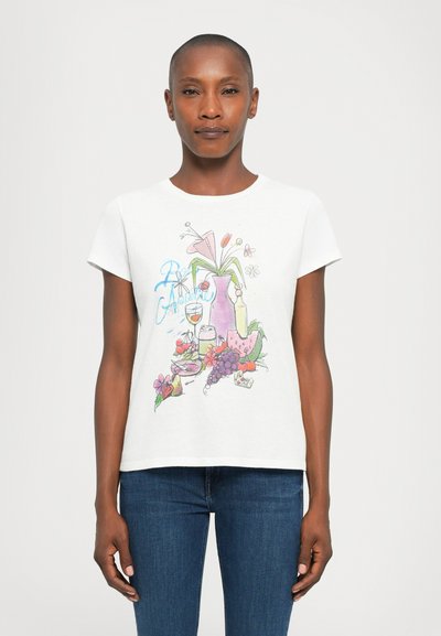Woman wearing a white T-shirt with a colorful print of flowers, fruits, a vase, and a glass, paired with blue jeans, standing neutral pose.