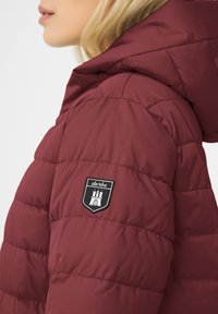 Burgundy puffer jacket with a hood. Features quilted sections and a black shield logo on the left arm. Smooth texture, durable fabric.