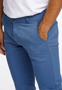 Blå bomullshängselbyxor med smal passform, sidofickor och en mjuk, lätt textur. Inkluderar dragkedja och knappstängning.