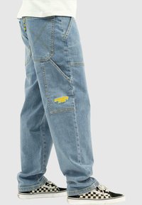 Lysblå baggy denimbukser med flere lommer og kontrasterende syninger. Har et gult "homeboy xtra" logo og ternede sneakers.