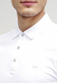 Weißes Polohemd mit Kragen, drei Knopf-Leiste und silbernem Faden-Logo auf der Brust. Glatte Textur, lässiges Design.