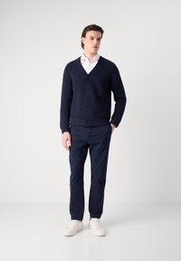Marine gebreide cardigan met een V-hals, vier knopen en een gestructureerde stof, gecombineerd met marine chinos en lichte sneakers. Achtergrond is egaal wit.