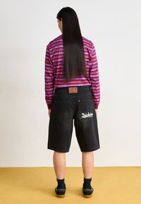 Shorts en denim noir avec une coupe ample, dotés d'un patch logo au dos et de deux poches arrière. Le modèle porte une chemise à rayures, violette et rose, à manches longues.