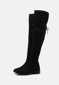 Botas negras por encima de la rodilla hechas de suave ante. Presentan una punta redonda, suela de goma plana y un detalle de lazo en la parte posterior.