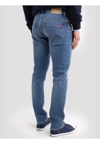 Mees, kes kannab helesiniseid teksapükse ja tumesiniseid tossusid, seisab seljaga vaataja poole lihtsa tausta ees.