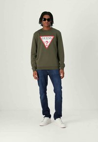 Νεαρός άνδρας με μαύρα γυαλιά ηλίου, ελαιοπράσινη μπλούζα με κόκκινο και λευκό λογότυπο "GUESS", σκούρα μπλε τζιν και άσπρα αθλητικά παπούτσια, που στέκεται μπροστά από ένα απλό φόντο.